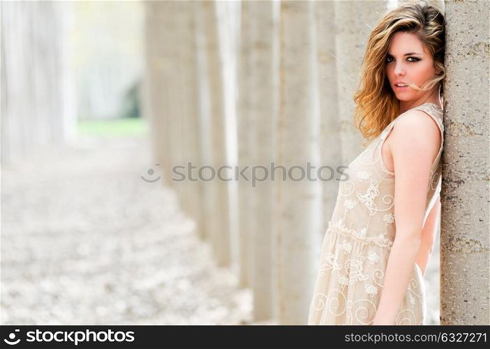 Beautiful blonde girl, dressed with a beige dress, standing in a poplar forest