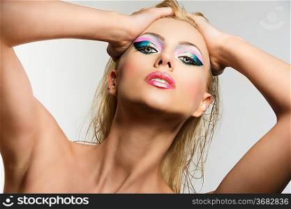 beautiful blonde girl with showy make up, her hands are on her head and she looks in to the lens with aggressive expression