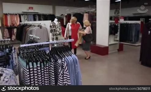 Beautiful blonde ladies with shopping bags choosing clothes in fashion boutique store. Excited attractive elderly woman trying on elegant red dress and showing to her firend while shopping together in garments shop. Slow motion.