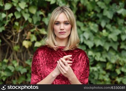 Beautiful blonde woman in green leaves background. Young girl wearing red dress standing in the street. Pretty female with straight hairstyle and blue eyes.