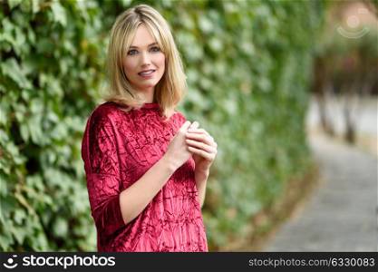 Beautiful blonde woman in green leaves background. Young girl wearing red dress standing in the street. Pretty female with frizzy hairstyle and blue eyes.