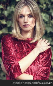 Beautiful blonde woman in green leaves background. Young girl wearing red dress standing in the street. Pretty female with frizzy hairstyle and blue eyes.