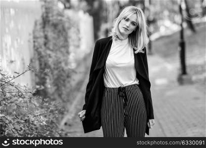 Beautiful blonde woman in urban background. Young girl wearing black blazer jacket and striped trousers standing in the street. Pretty female with straight hair hairstyle and blue eyes.