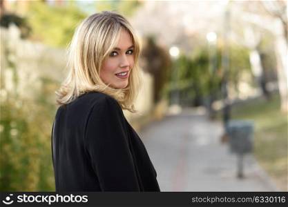Beautiful blonde woman in urban background. Young girl wearing black blazer jacket standing in the street. Pretty female with straight hair hairstyle and blue eyes.