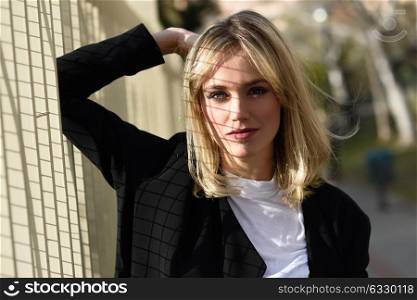 Beautiful blonde woman in urban background. Young girl wearing black blazer jacket standing in the street. Pretty female with straight hair hairstyle and blue eyes.