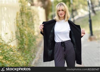 Beautiful blonde woman in urban background. Young girl wearing black blazer jacket and striped trousers standing in the street. Pretty female with straight hair hairstyle and blue eyes.