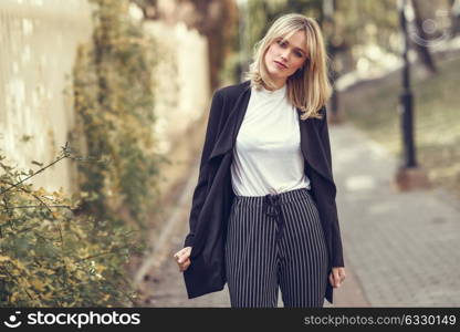 Beautiful blonde woman in urban background. Young girl wearing black blazer jacket and striped trousers standing in the street. Pretty female with straight hair hairstyle and blue eyes.