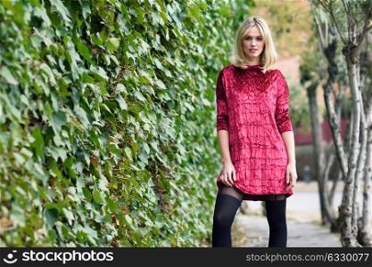 Beautiful blonde woman in urban background. Young girl wearing red dress and tights standing in the street. Pretty female with straight hairstyle and blue eyes.