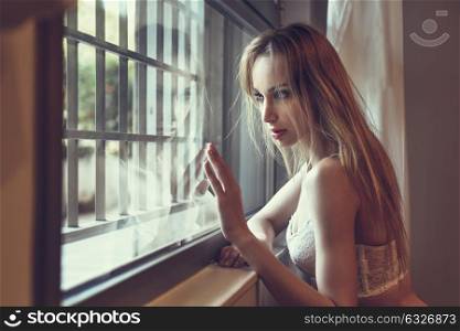 Beautiful blonde woman in white lingerie posing near a window. Young girl wearing underwear with long hair.