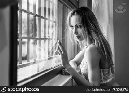 Beautiful blonde woman in white lingerie posing near a window. Young girl wearing underwear with long hair. Black and white photograph.