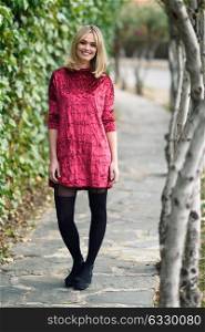 Beautiful blonde woman smiling in urban background. Young girl wearing red dress and tights standing in the street. Pretty female with straight hairstyle and blue eyes.