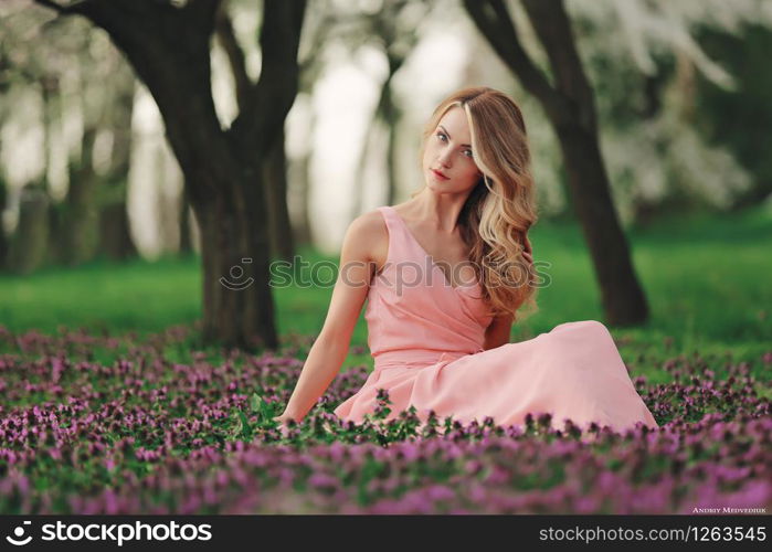 Beautiful blonde young woman in colorful flowers. girl with make-up and hairstyle in pink dress in blossoming spring park. Art work of romantic woman .Pretty tenderness model looking at the camera.. Beautiful blonde young woman in colorful flowers. girl with make-up and hairstyle in pink dress in blossoming spring park. Art work of romantic woman. Pretty tenderness model looking at the camera