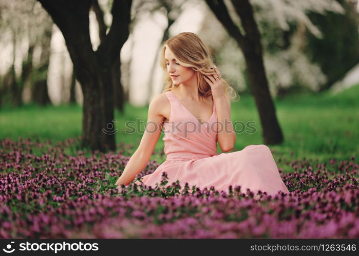 Beautiful blonde young woman in colorful flowers. girl with make-up and hairstyle in pink dress in blossoming spring park. Art work of romantic woman .Pretty tenderness model looking at the camera.. Beautiful blonde young woman in colorful flowers. girl with make-up and hairstyle in pink dress in blossoming spring park. Art work of romantic woman. Pretty tenderness model looking at the camera