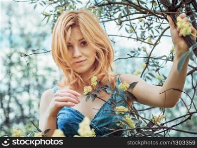 Beautiful blonde young woman in denim topic gently smiling with sympathy looking at the camera, standing among flowering branches on nature. Selective focus, blurred vignette.