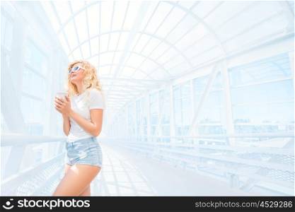 Beautiful blonde young woman in sunglasses with take away coffee cup standing on the bridge.