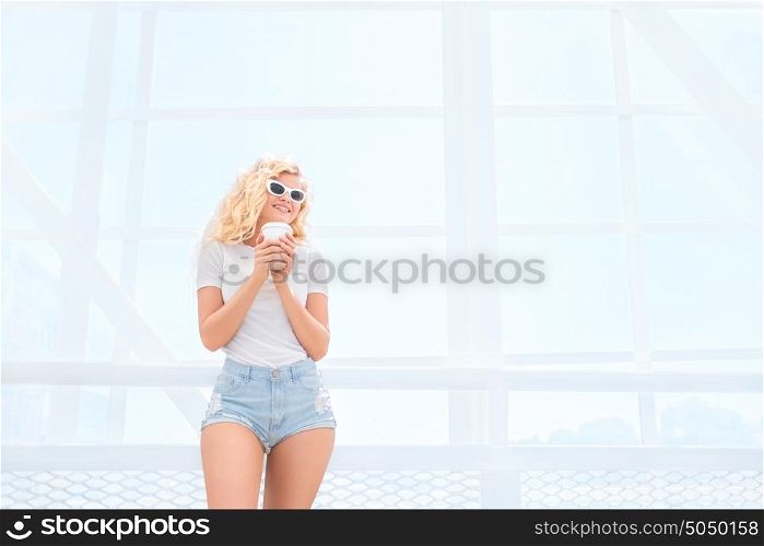 Beautiful blonde young woman in sunglasses with take away coffee cup standing on the bridge.