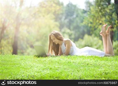 Beautiful Blonde Young Woman Lying with Laptop on the Beautiful Green Meadow in the Bright Sunny Day