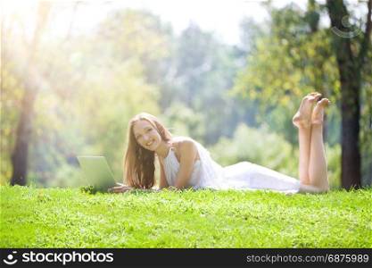 Beautiful Blonde Young Woman Lying with Laptop on the Beautiful Green Meadow in the Bright Sunny Day