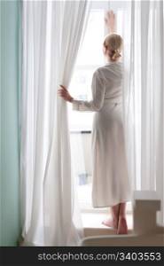 Beautiful blonde young woman, silk bathrobe, standing by the window, between the curtains, natural light