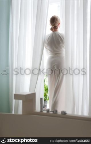 Beautiful blonde young woman, silk bathrobe, standing by the window, natural light