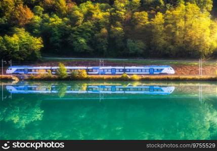 Beautiful blue modern high speed train and river in alpine mountains at sunrise in autumn. Passenger train, reflection in water, railroad, lake, orange trees in fall. Railway station in Slovenia