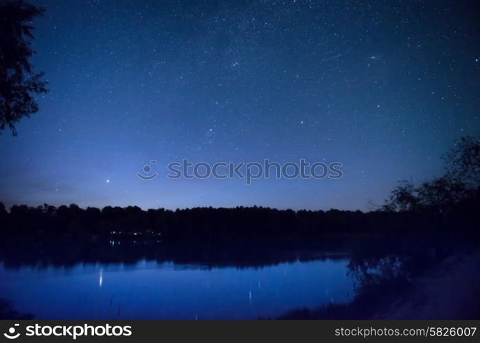 Beautiful blue night sky with many stars on a lake with forest on the other coast. Milkyway reflection in water