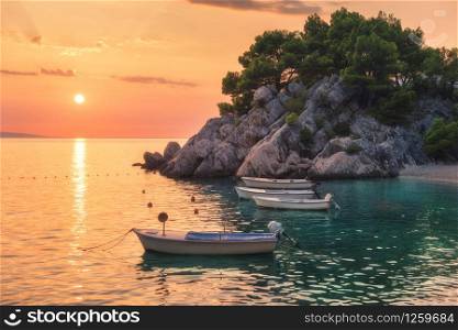 Beautiful boats on the sea and green trees growing out of the rock at sunset in summer in Croatia. Colorful landscape with motorboats, sandy beach, cliff and orange sky. Travel. Nature background