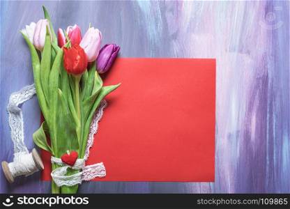 Beautiful bouquet of colorful tulips tied with white lace ribbon and a wooden clip with a red heart, placed on a blank red paper sheet.
