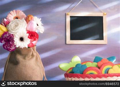 Beautiful bouquet of diverse flowers in a jute sack, a basket with multi-shaped colorful cookies and a blank blackboard hanging on the purple wall.