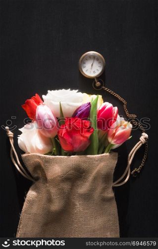 Beautiful bouquet of diverse flowers wrapped in a rustic jute sack and a vintage pocket clock, on a black background, on a sunny day of spring.