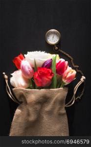 Beautiful bouquet of diverse flowers wrapped in a rustic jute sack and a vintage pocket clock, on a black background, on a sunny day of spring.