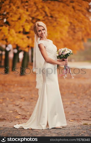 Beautiful bride in luxury white long dress with wedding bouquet in autumn park on wedding day.. Beautiful bride in luxury white long dress with wedding bouquet in autumn park on wedding day