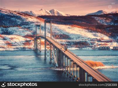 Beautiful bridge at sunset in Lofoten islands, Norway. Aerial winter landscape with cars on the road, blue sea, forest, snowy mountains, colorful orange sky with clouds. Nordic scenery. Travel