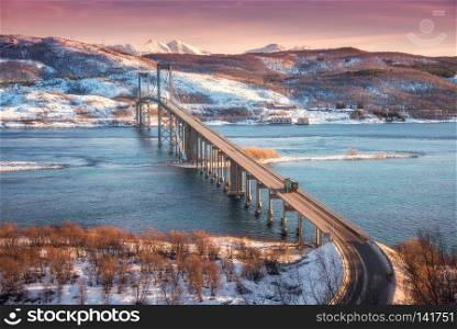 Beautiful bridge during sunset in Lofoten islands, Norway. Aerial winter landscape with cars on the road, blue sea, trees, snowy mountains, colorful red sky with clouds, buildings. Nordic scenery