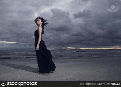 Beautiful brunete model in long black dress standing on the beach. Cloudy weather. Sunset