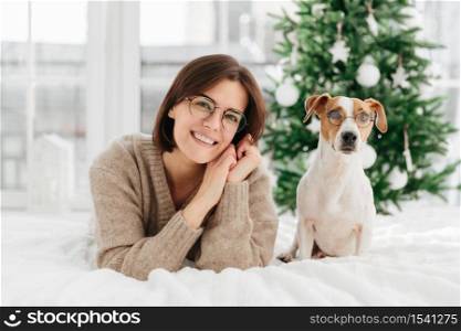 Beautiful brunette adult woman wears big round spectacles, tilts head, wears warm winter sweater, her pet in glasses poses near at bed, express happy emotions, await for Christmas, enjoy coziness