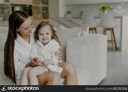 Beautiful brunette affectionate mother holds small curly haired cute dauhter in dressing gown pose in cozy room against modern apartment interior. Mom and little girl at home after taking shower