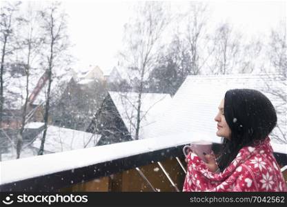 Beautiful brunette woman covered with a red blanket, holding a cup of hot drink, enjoying the snowfall with her eyes closed, on a balcony.