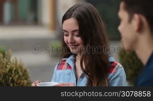 Beautiful brunette woman drinking hot cup of coffee or tea, enjoying its taste and aroma while having romantic date in open air coffee shop. Charming girl sipping hot drink from white cup at outdoors cafe and talking to her boyfriend.