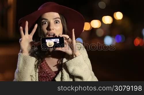 Beautiful brunette woman in bordo hat showing mobile phone with special offer, halloween sale on screen in night city street. Emotional girl gesturing, making funny facial expressions and holding mobile phone with discount text on screen at night.