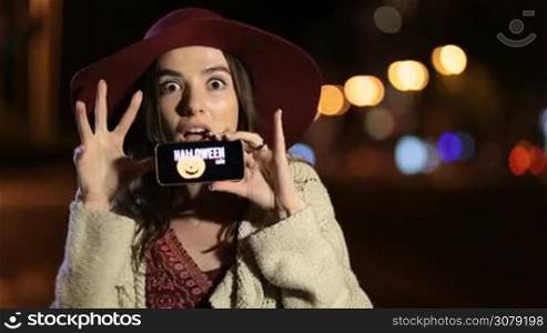 Beautiful brunette woman in bordo hat showing mobile phone with special offer, halloween sale on screen in night city street. Emotional girl gesturing, making funny facial expressions and holding mobile phone with discount text on screen at night.