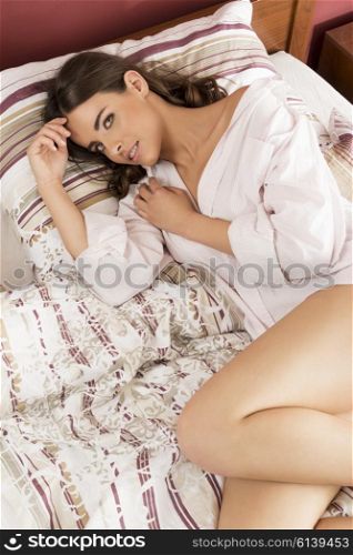 beautiful brunette woman lying relaxed on comfortable bed with nude legs and unbuttoned shirt. She is looking in camera with happy expression