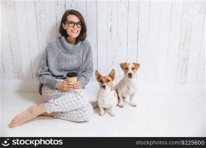 Beautiful brunette woman sits on floor with her two favourite dogs, dressed casually, drinks takeaway coffee. Pleased female enjoys calm domestic atmosphere. People and animals, good relations