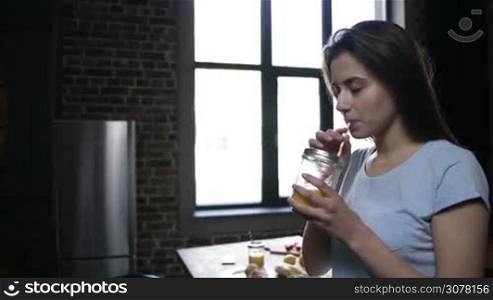 Beautiful brunette woman sitting on the table and drinking orange juice from mason jar in modern kitchen in the morning. Handsome man coming to his girlfriend and embracing her with love and passion. Slow motion. Side view. Steadicam stabilized shot.