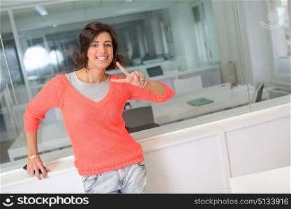Beautiful brunette woman smiling with casual clothes in an office. Girl making a v sign