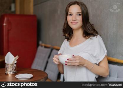 Beautiful brunette woman with dreamy expression, has coffee break after work, wears white t shirt, spends free time in coffee shop, enjoys recreation time. People, lifestyle and drinking concept