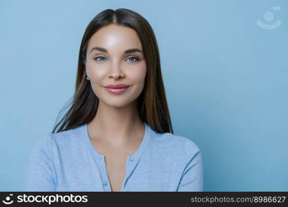 Beautiful brunette young woman has long dark hair, poses indoor against blue studio background, being successful businesswoman, has natural look, enjoys leisure or day off, stands delighted.