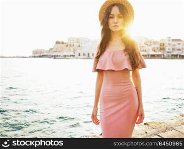 Beautiful brunette young woman wearing pink dress and straw hat enjoying sunrise on seafront in old european town. Fashion and style. Summer travel