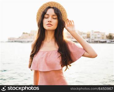 Beautiful brunette young woman wearing pink dress and straw hat enjoying sunrise on seafront in old european town. Fashion and style. Summer travel