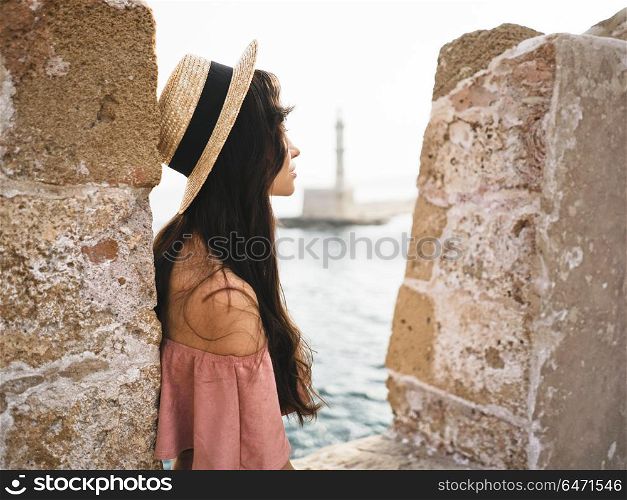 Beautiful brunette young woman wearing pink dress and straw hat enjoying sunrise on seafront in old european town. Fashion and style. Summer travel. Stylish brunette woman enjoying sunrise on seafront